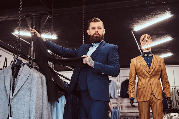 Free photo elegantly dressed bearded male with tattoos on hands and neck chooses new suit in a menswear store.