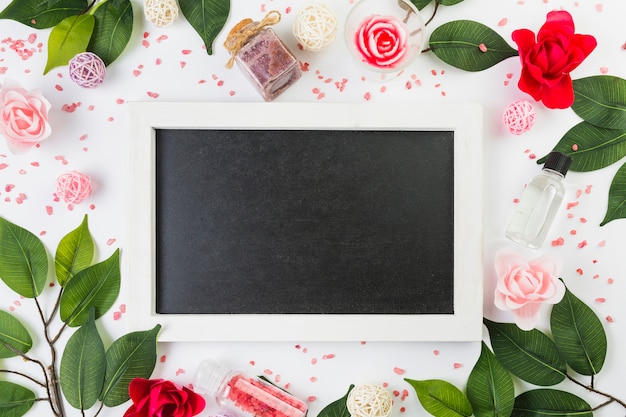 Free photo elevated view of blank frame surrounded with cosmetic products and leaves on white surface