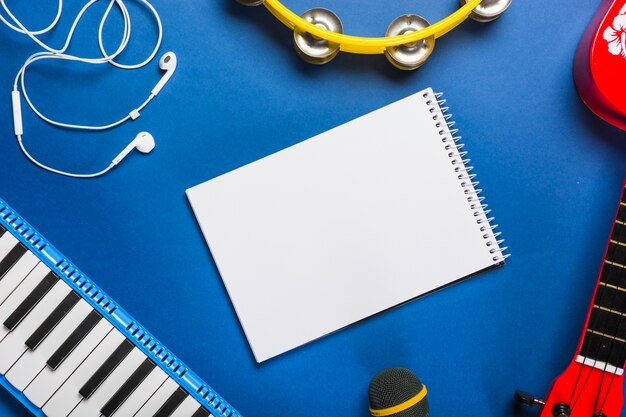 Free Photo elevated view of blank spiral notepad surrounded with earphone; guitar; microphone; piano keyboard and tambourine over blue backdrop