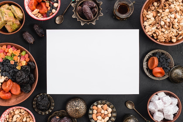 An elevated view of blank white paper surrounded with delicious dried fruits; nuts and sweets for ramadan on black textured backdrop