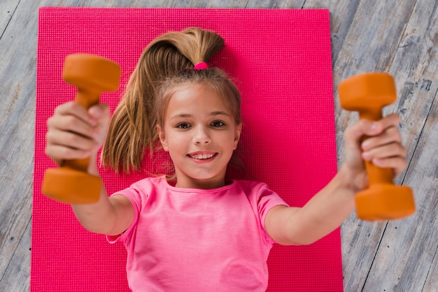 An elevated view of a blonde girl lying on pink carpet exercising with dumbbell