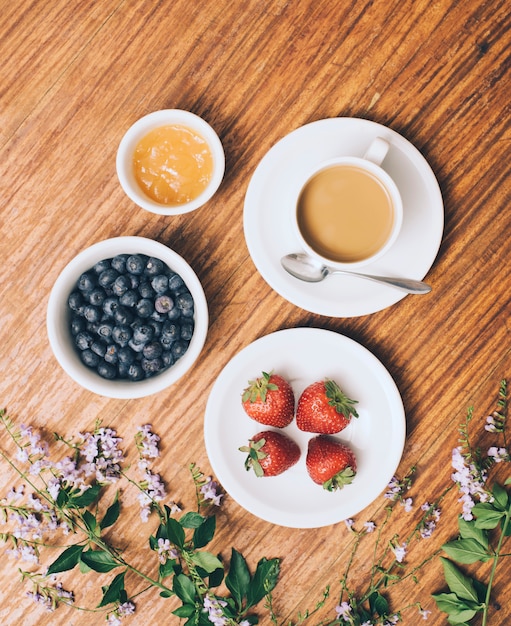 An elevated view of blueberry; jam; strawberry and coffee cup on flowers against wooden backdrop