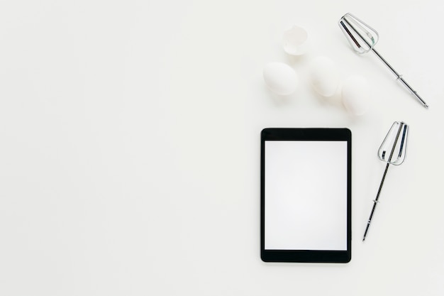 Elevated view of digital tablet with whisk and eggs on white background