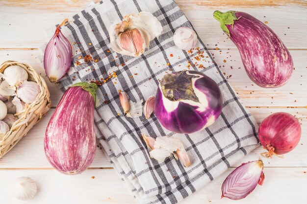 Elevated view of eggplants; onion; garlic cloves and chequered pattern cloth on wooden plank