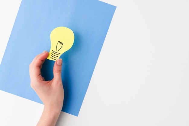 An elevated view of female hand holding light bulb paper cutout over blue card paper