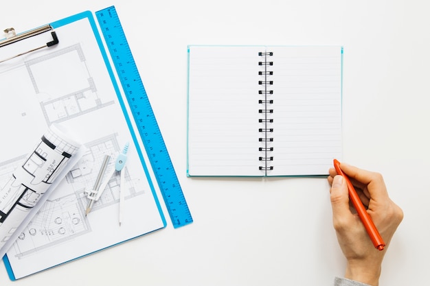 Elevated view of human hand writing on diary near blueprint clipboard