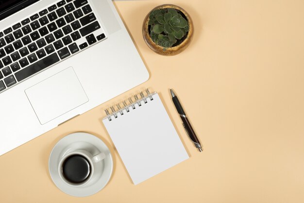 Elevated view of laptop; coffee cup; pen; and spiral notepad over beige background