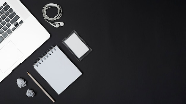 Elevated view of laptop; earphones; crumpled papers; pencil; blank spiral notepad and frame on black background