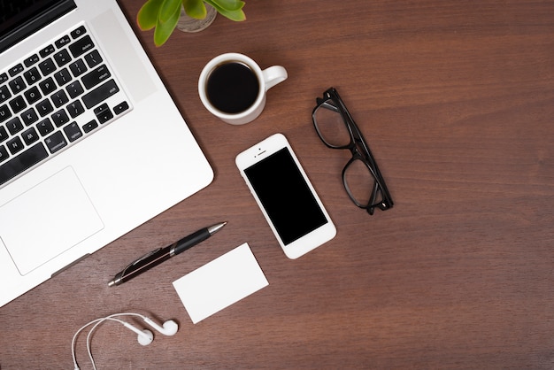 Elevated view of laptop; mobile phone; tea; earphones; pen and eye glasses on wooden table