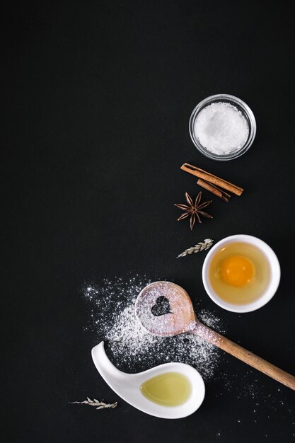 Elevated view of oil; egg; flour; sugar; grains; spices and heart shape wooden spoon on black surface