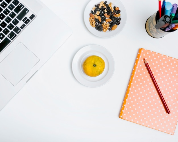 Elevated view of pencil; citrus fruit; notebook; nut food and laptop on white backdrop