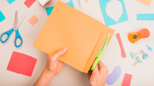 Elevated view of person's hand cutting paper with zigzag scissor with craft accessories on white table