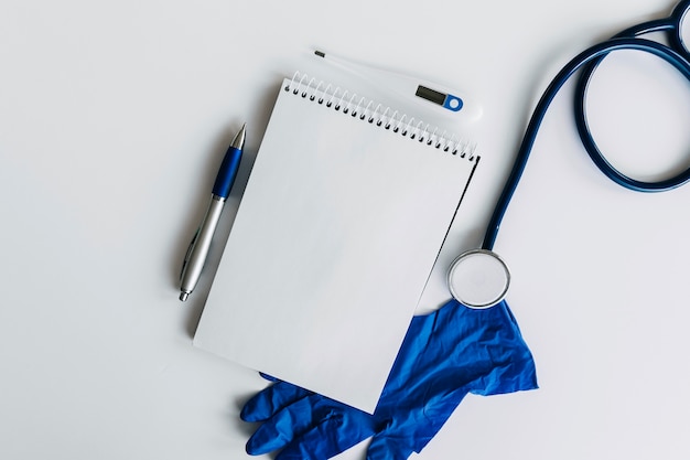 Free photo elevated view of spiral notepad; pen; stethoscope; thermometer and gloves on white backdrop