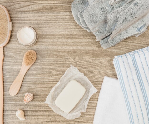 Elevated view of sponge; seashell; soap; brush; towel and moisturizing cream on wooden background