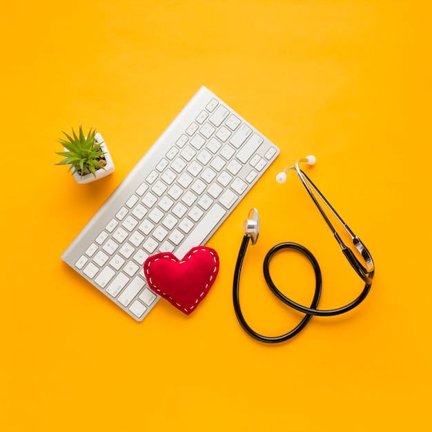 Free photo elevated view of stethoscope; stitched heart shape; wireless keyboard; succulent plant over yellow backdrop