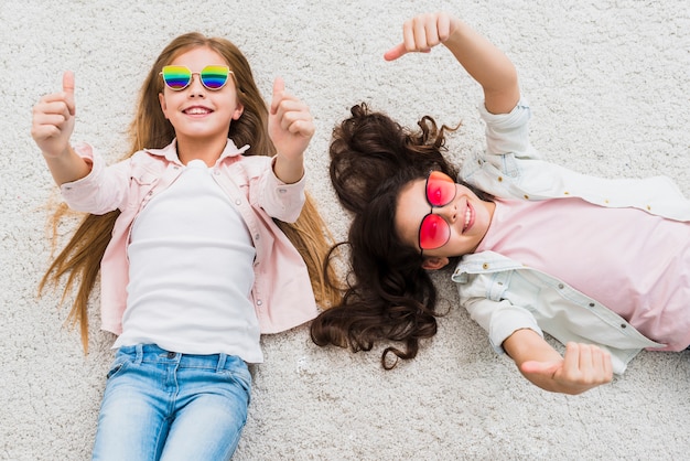 Free photo an elevated view of two female friends lying on carpet showing thumb up sign