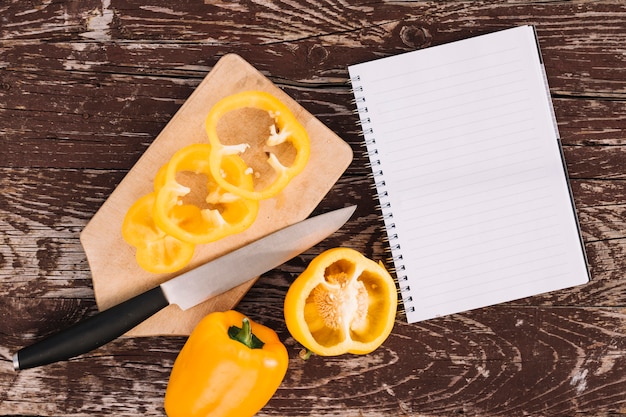 An elevated view of yellow bell pepper on chopping board with knife and spiral notebook on wooden desk