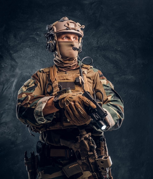 Free photo elite unit, special forces soldier in camouflage uniform holding a gun with a flashlight and looking sideways. studio photo against a dark textured wall