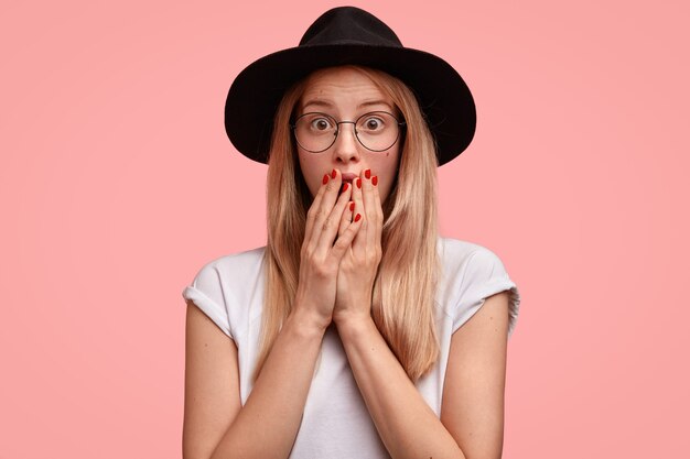 Embarrassed beautiful woman with blonde hair, covers mouth with both hands, has red manicure, feels shocked as sees something awful