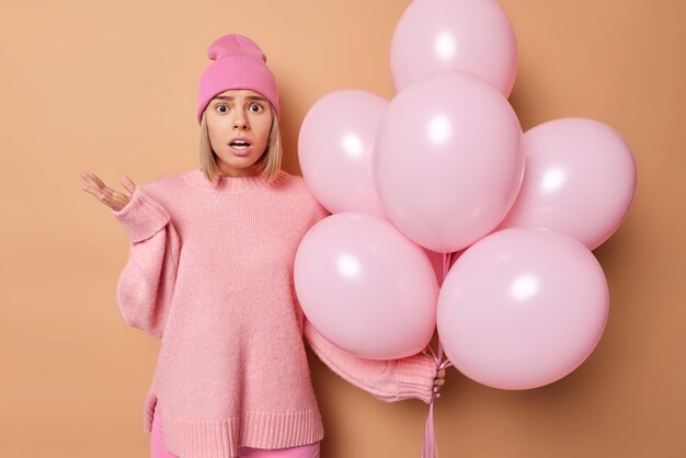 Free Photo embarrassed blonde young woman wears casual jumper and hat poses with helium balloons shocked to hear bad news celebrates anniversary isolated over brown background. birthday celebration concept