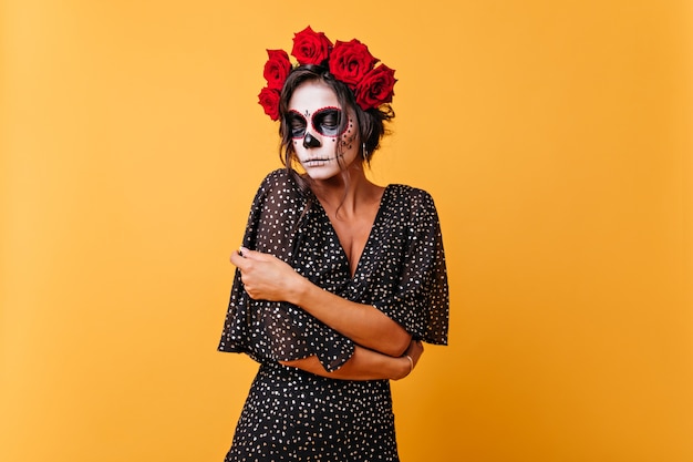 Free photo embarrassed dark-haired girl with flowers on her head posing in traditional calavera skull. portrait of model from mexico who looked down to the floor