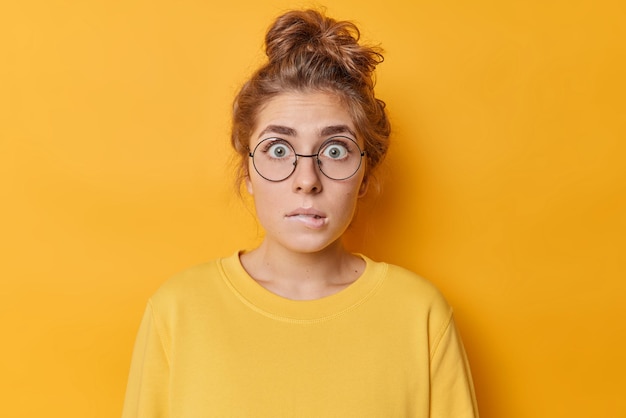 Embarrassed nervous woman with anxious expression bites lips stars through round spectacles finds out something dressed in casual jumper isolated over yellow background. Worried female model