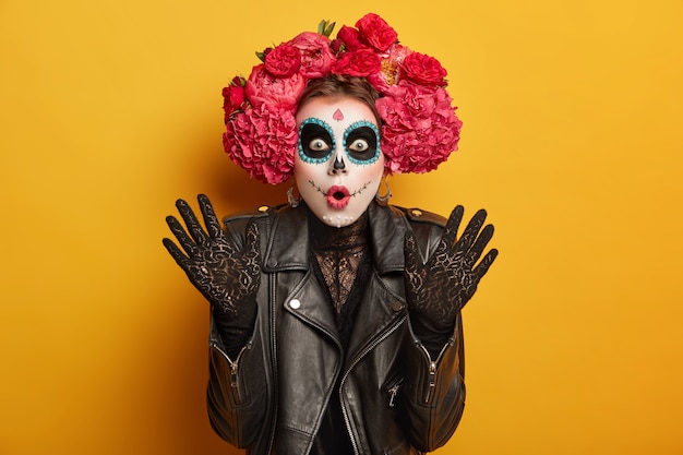 Free photo embarrassed shocked woman dressed in traditional clothing or attire to honor dead in mexico, has widely opened mouth, skull makeup, raises palms from amazement