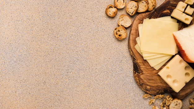 Emmental cheese and gouda cheese with slices on coaster with bread slices and walnut over beige textured backdrop