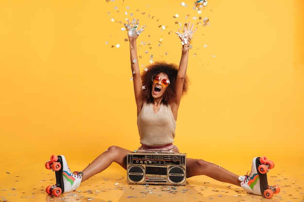 Free photo emotional african disko woman in retro wear and roller scates throwing confetti while sitting with boombox