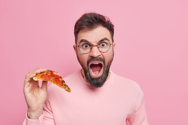 Free photo emotional bearded adult man screams loudly holds slice of tasty appetizing pizza eats fast food for snack dressed in casual clothes