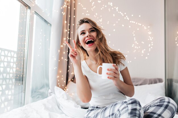 Emotional blonde girl drinking coffee and fooling around. Romantic lady with cup of tea posing with peace sign.