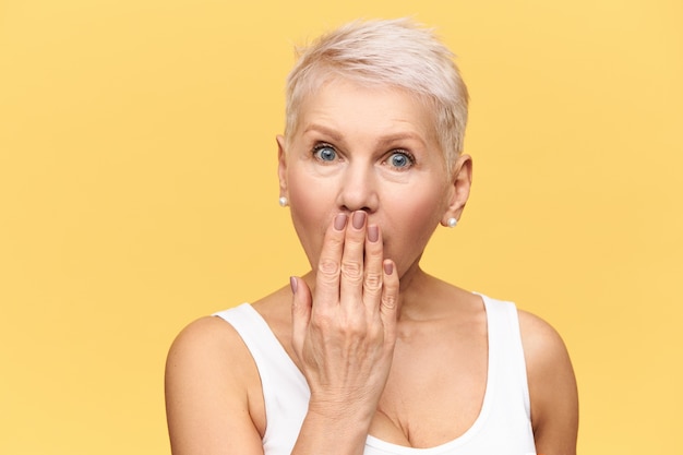  emotional blonde middle aged woman widening eyes and covering mouth with hand, trying not to tell intriguing information or secret, having surprised astonished facial expression