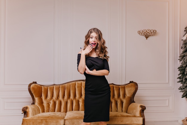 Emotional, bright brown-haired woman in black dress surprisingly looks and touches corner of her lips