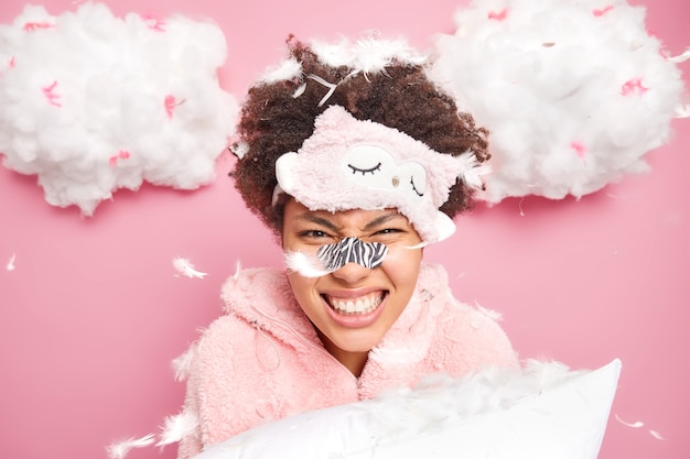 Emotional curly woman clenches teeth feels angry after pillow fight poses under feathers flying around wears sleepmask applies patch on nose to reduce blackheads isolated over pink wall