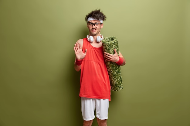 Free photo emotional displeased man makes stop gesture, asks not to bother him, holds rolled up karemat, stays in good physical shape, going to have workout in gym, poses agaist green wall. sport concept