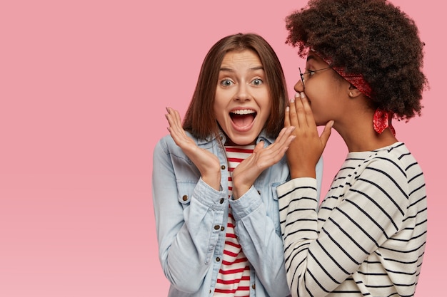 Free photo emotional european woman clasps hands and exclaims loudly as hears rumors from best friend. afro american woman whispers secret at companions ear