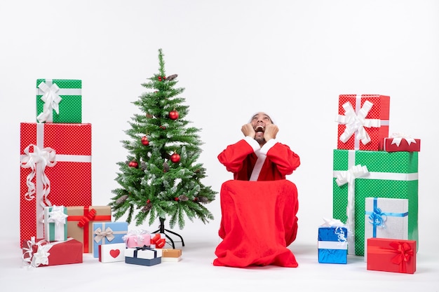 Free Photo emotional excited concerned young man dressed as santa claus with gifts and decorated christmas tree on white background stock photo