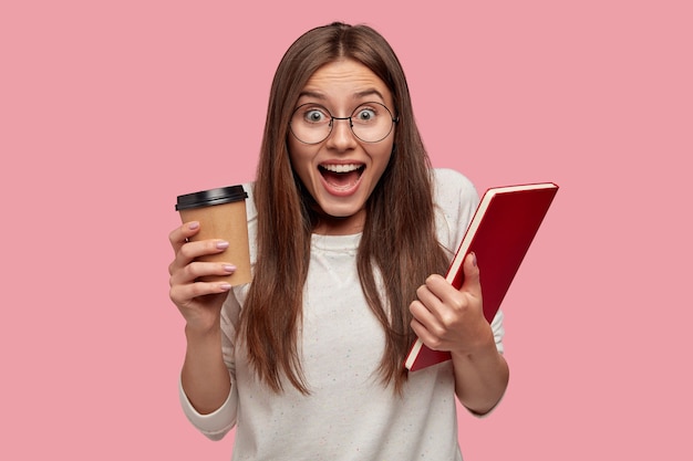 Emotional excited young lady with dark hair, shouts with happiness, carries takeaway coffee and book, dressed in white jumper, feels overjoyed, isolated over pink wall. Emotions concept