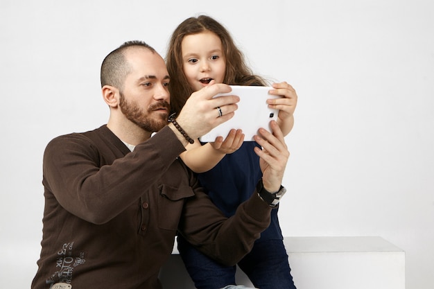 Emotional female child opening mouth widely, amazed with new digital tablet, using it with her fashionable modern young father with stubble