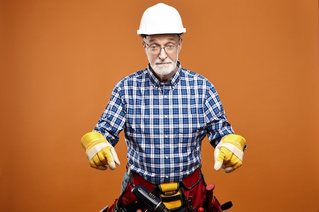 Emotional funny elderly senior house builder wearing protective helmet, safety gloves and bag with instruments around his waist having shocked surprised facial expression, pointing fore fingers down