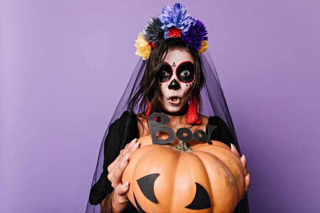 Emotional girl shouts "boo" holding huge decorated pumpkin in front of her. Portrait of black bride painted in shape of skull.