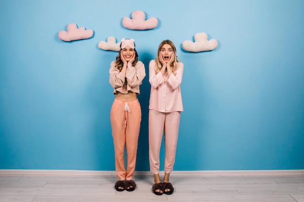 Free photo emotional girls posing in pink pajamas studio shot of two laughing friends standing on blue background with fake clouds