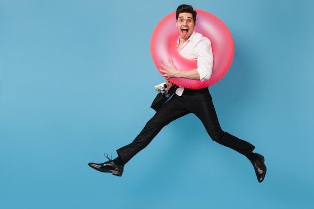 Free photo emotional guy in pants and white shirt joyfully looks into camera and jumps with inflatable circle on blue background