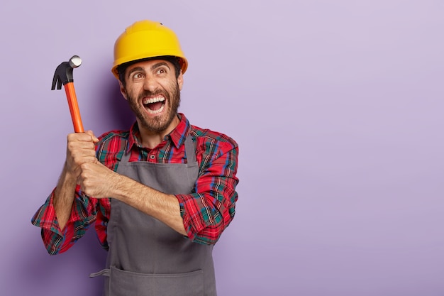 Emotional handyman holds hammer with both hands, ready for labour work, wears yellow protective helmet, casual workwear, yells emotionally