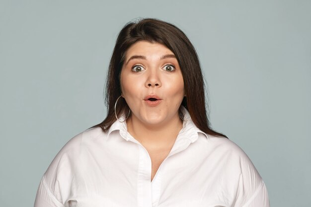 Emotional obese chubby young brunette female employee in formal shirt opening mouth expressing full disbelief, receiving unexpected raise in salary. Human facial expressions, feelings and reaction