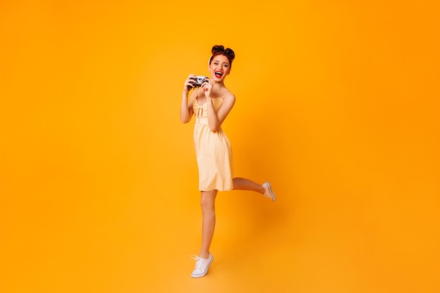 Emotional pinup girl with camera dancing on yellow background Studio shot of female photographer in dress