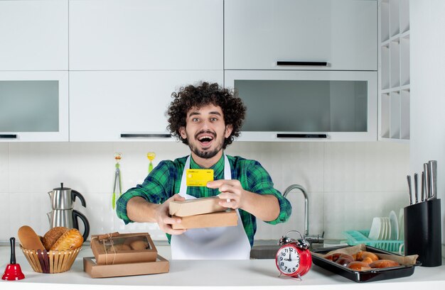 Emotional positive young man standing behind the table various pastries on it and holding bank card brown boxes in the white kitchen