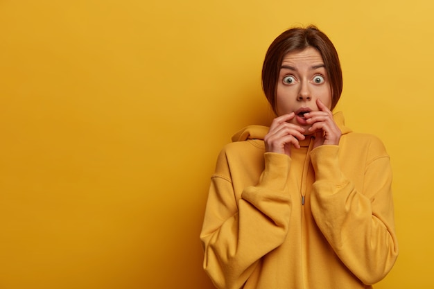 Emotional scared anxious concerned woman looks with great fear at camera, cannot believe her eyes, holds breath, wears yellow sweatshirt