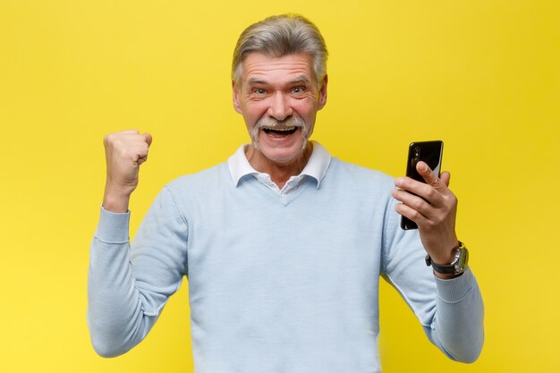 Emotional senior man with phone winn something while posing on yellow wall