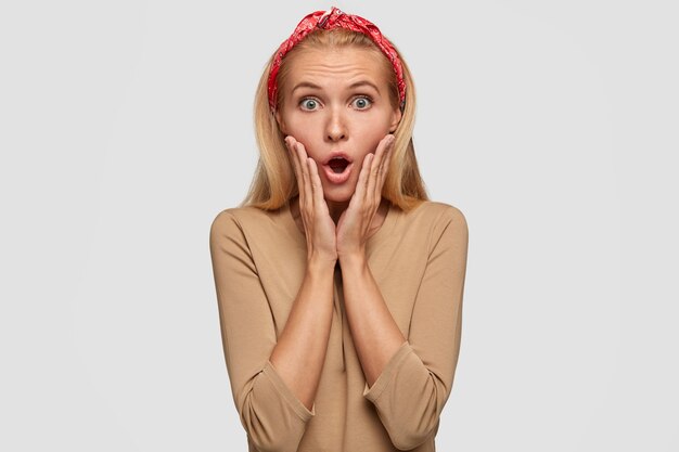 Emotional surprised blonde woman touches cheeks with palms, wears red headband and beige sweater